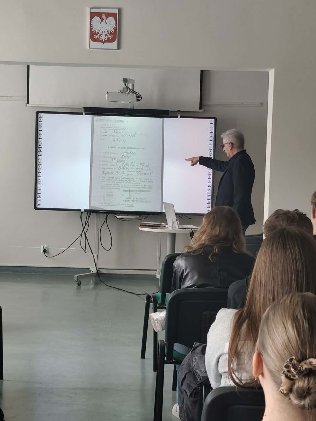 uczniowie siedzący na krzesłach słuchają wypowiedzi mężczyzny