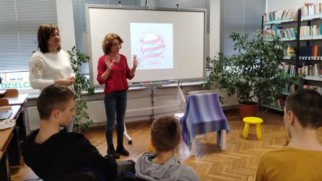 Uczniowie siedzą na krzesłach nauczyciele stoją i prowadzą zajęcia.